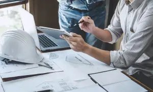 Inżynierowie analizują plany, by ustalić, jak wygląda pozwolenie na budowę. Na stole biały kask, laptop i dokumenty.
