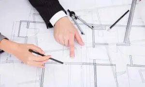 Rysunek techniczny z zaznaczonymi zmianami w projekcie budowlanym. Dłonie wskazują na plany, obok cyrkiel i ołówek.