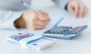 Ręce piszą przy kalkulatorze, obok stos banknotów 500 euro. Analiza finansowa, w tym euribor 3m.