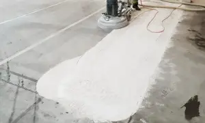 Maszyna szlifuje beton, tworząc biały pył. Widać ślady butów na powierzchni, co sugeruje, że źle zawibrowany beton wymaga poprawy.