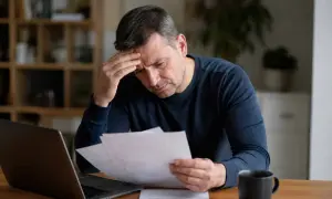 Zmartwiony mężczyzna z ręką na czole analizuje dokumenty, być może związane z regres ubezpieczeniowy. Obok laptop i kubek.