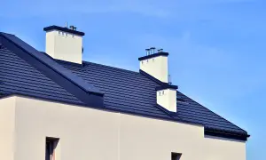 Nowoczesny dach bez okapu z ciemnogranatową dachówką i białymi kominami na tle błękitnego nieba.