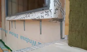 Montaż okien w domu szkieletowym. Widać metalowe wsporniki pod parapetem, membranę wiatroizolacyjną i izolację.