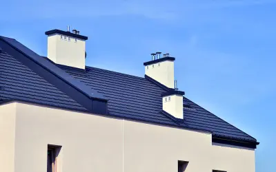 Nowoczesny dach bez okapu z ciemnogranatową dachówką i białymi kominami na tle błękitnego nieba.
