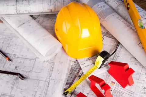 Plany budowlane, żółty kask, miarka i poziomica – to wszystko elementy metody gospodarczej w budownictwie.