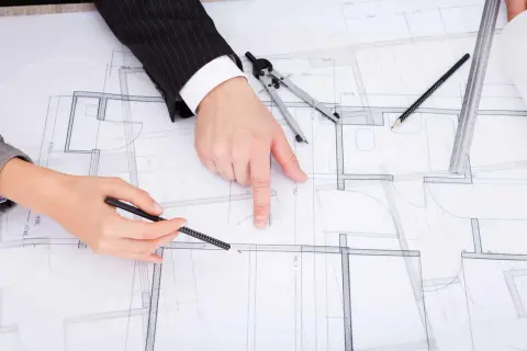 Rysunek techniczny z zaznaczonymi zmianami w projekcie budowlanym. Dłonie wskazują na plany, obok cyrkiel i ołówek.