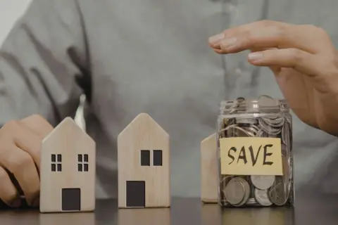 Dwa drewniane domki i słoik z monetami z napisem "SAVE". Czy mieszkanie może być wkładem własnym?