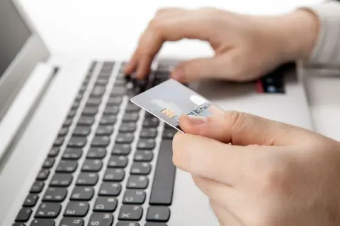 Dłoń trzyma kartę płatniczą nad klawiaturą laptopa. Czy nazwa odbiorcy w przelewie ma znaczenie?