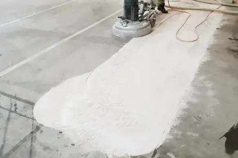 Maszyna szlifuje beton, tworząc biały pył. Widać ślady butów na powierzchni, co sugeruje, że źle zawibrowany beton wymaga poprawy.