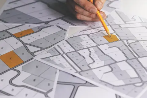 Rysunek geodezyjny z zaznaczonymi działkami. Dłoń z ołówkiem planuje scalenie działek, analizując mapę z numeracją.