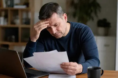 Zmartwiony mężczyzna z ręką na czole analizuje dokumenty, być może związane z regres ubezpieczeniowy. Obok laptop i kubek.