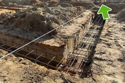 Wykonano wykopy pod fundamenty, w których widać zbrojenie. W tle plac budowy z hałdami ziemi i elementami konstrukcji. Widać ławy ciesielskie.