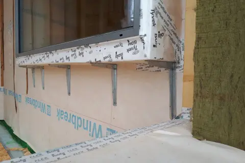 Montaż okien w domu szkieletowym. Widać metalowe wsporniki pod parapetem, membranę wiatroizolacyjną i izolację.