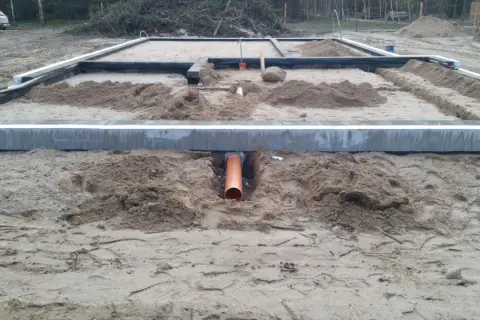 Pomarańczowa rura kanalizacyjna przechodzi przez wykop pod fundamentem, zapewniając przepust wody.