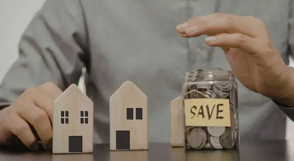 Dwa drewniane domki i słoik z monetami z napisem "SAVE". Czy mieszkanie może być wkładem własnym?