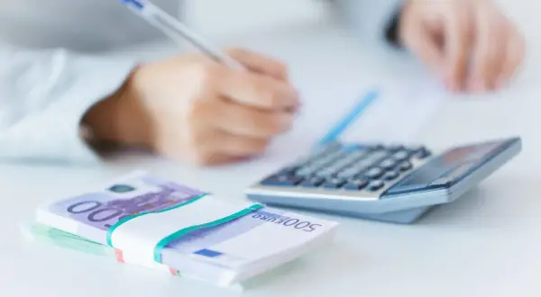 Ręce piszą przy kalkulatorze, obok stos banknotów 500 euro. Analiza finansowa, w tym euribor 3m.