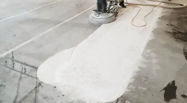 Maszyna szlifuje beton, tworząc biały pył. Widać ślady butów na powierzchni, co sugeruje, że źle zawibrowany beton wymaga poprawy.