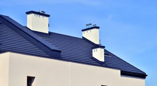 Nowoczesny dach bez okapu z ciemnogranatową dachówką i białymi kominami na tle błękitnego nieba.