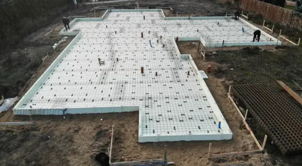 Prace na działce budowlanej: fundament z płytami izolacyjnymi i zbrojeniem, gotowy do zalania betonem. Glina wokół placu budowy.