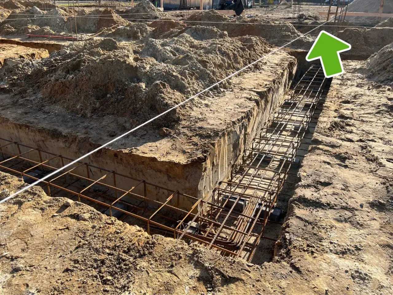 Wykonano wykopy pod fundamenty, w których widać zbrojenie. W tle plac budowy z hałdami ziemi i elementami konstrukcji. Widać ławy ciesielskie.