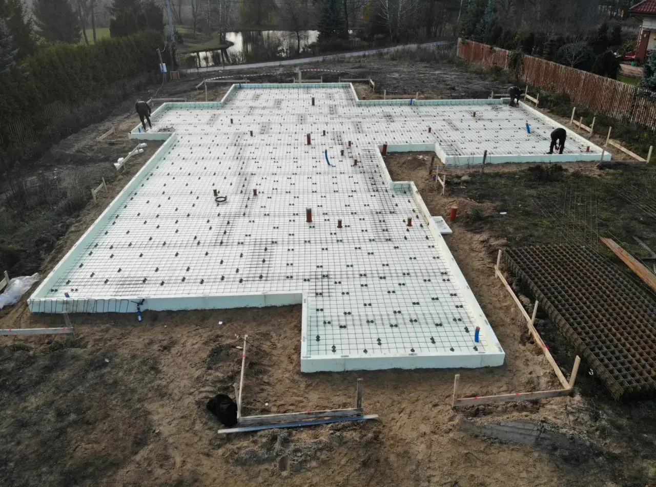 Prace na działce budowlanej: fundament z płytami izolacyjnymi i zbrojeniem, gotowy do zalania betonem. Glina wokół placu budowy.