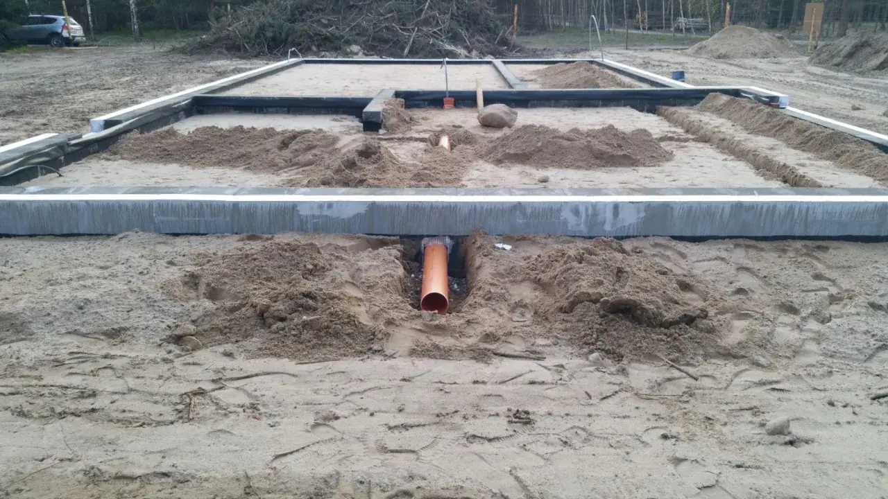Pomarańczowa rura kanalizacyjna przechodzi przez wykop pod fundamentem, zapewniając przepust wody.