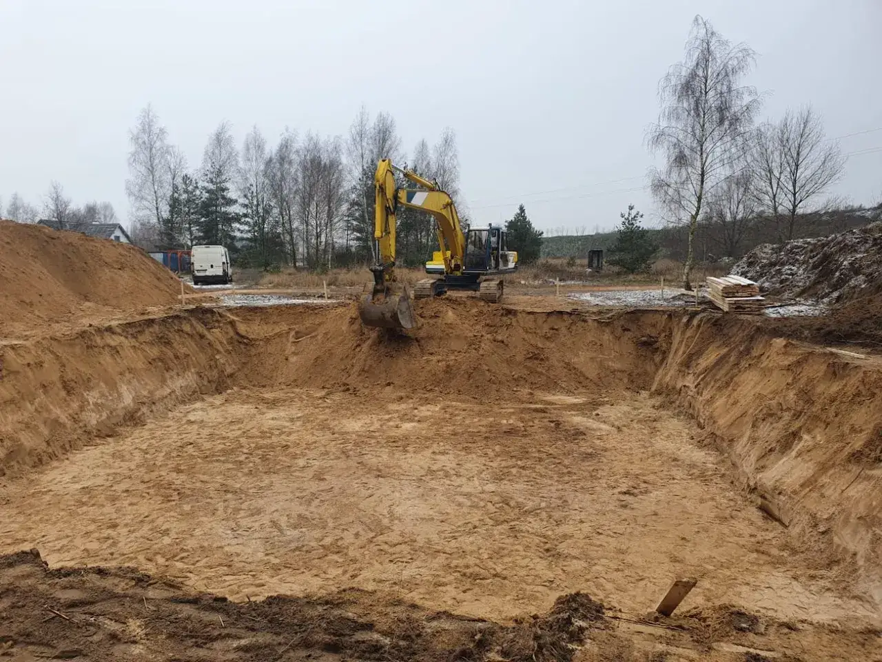 Ż&oacute;łty koparka wykonuje wykopy pod fundament. W tle teren budowy z hałdą piasku i drzewami.