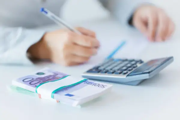 Ręce piszą przy kalkulatorze, obok stos banknotów 500 euro. Analiza finansowa, w tym euribor 3m.
