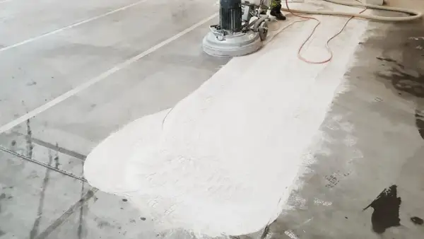 Maszyna szlifuje beton, tworząc biały pył. Widać ślady butów na powierzchni, co sugeruje, że źle zawibrowany beton wymaga poprawy.