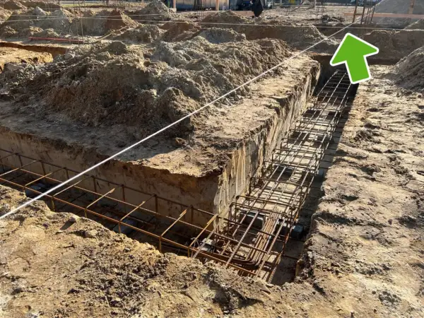 Wykonano wykopy pod fundamenty, w których widać zbrojenie. W tle plac budowy z hałdami ziemi i elementami konstrukcji. Widać ławy ciesielskie.