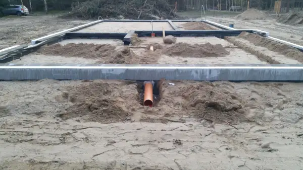 Pomarańczowa rura kanalizacyjna przechodzi przez wykop pod fundamentem, zapewniając przepust wody.
